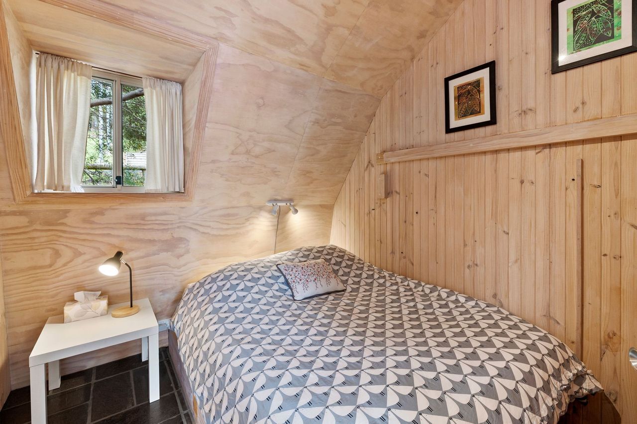 A bedroom with a bed, a bedside table with a lamp, a window with curtains, and wooden walls.