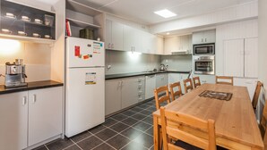 A modern kitchen with white cabinets, a built-in oven, a coffee machine, and a dining table with chairs. - Oceans 12 (Anglesea)