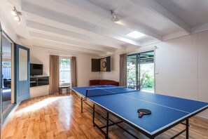 Ein geräumiger Raum mit einem Tischtennisplatte, einem Fernseher und Blick auf Grün hinter einer Schiebetür.