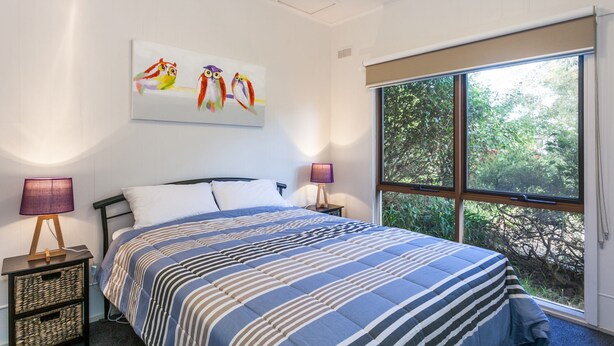 A bedroom with a large window, a bed with striped bedding, two bedside lamps, and a painting of owls on the wall. - Oceans 12 (Anglesea)