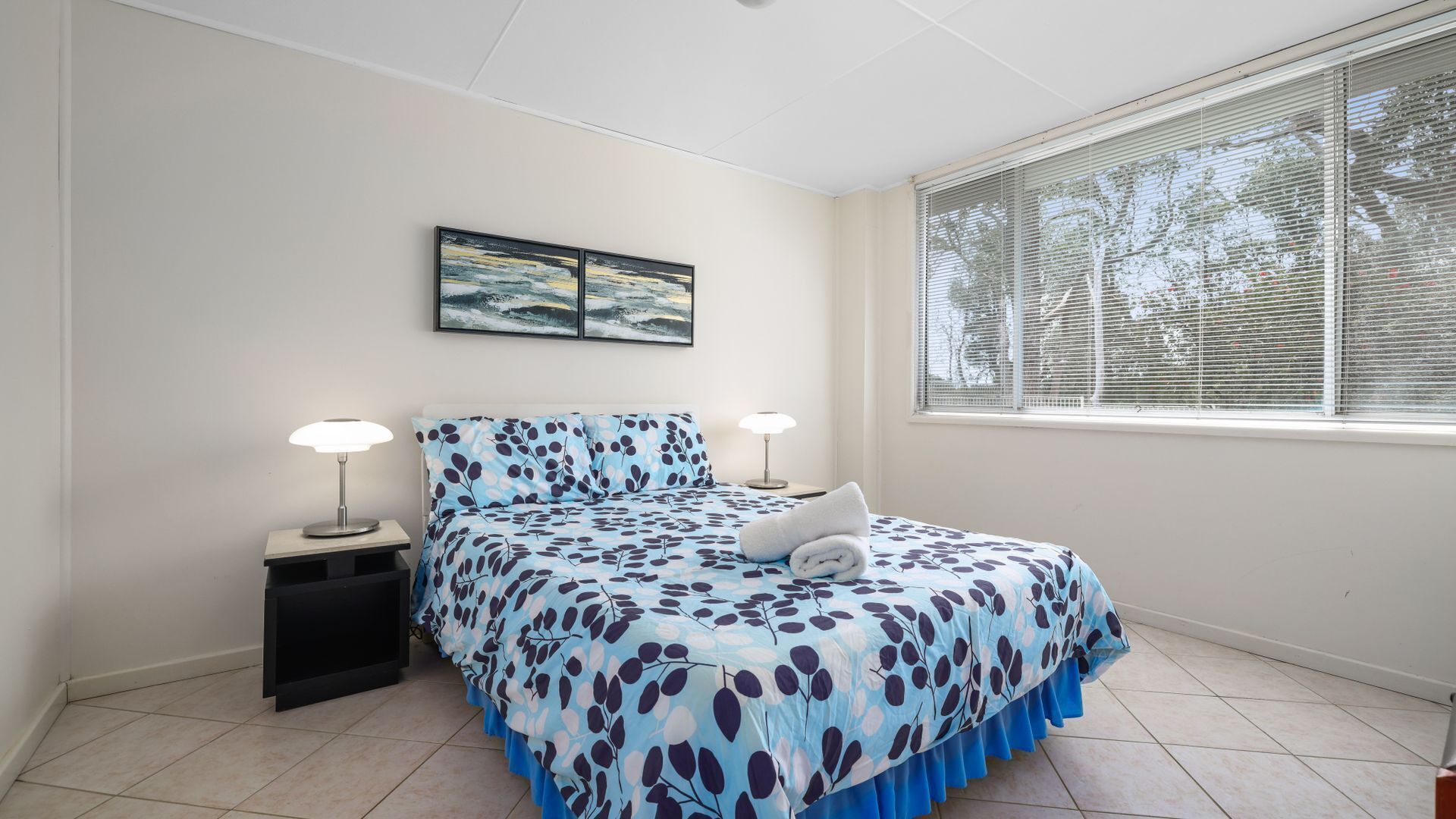 A bedroom with a bed, two bedside lamps, a window with blinds, and a painting on the wall.