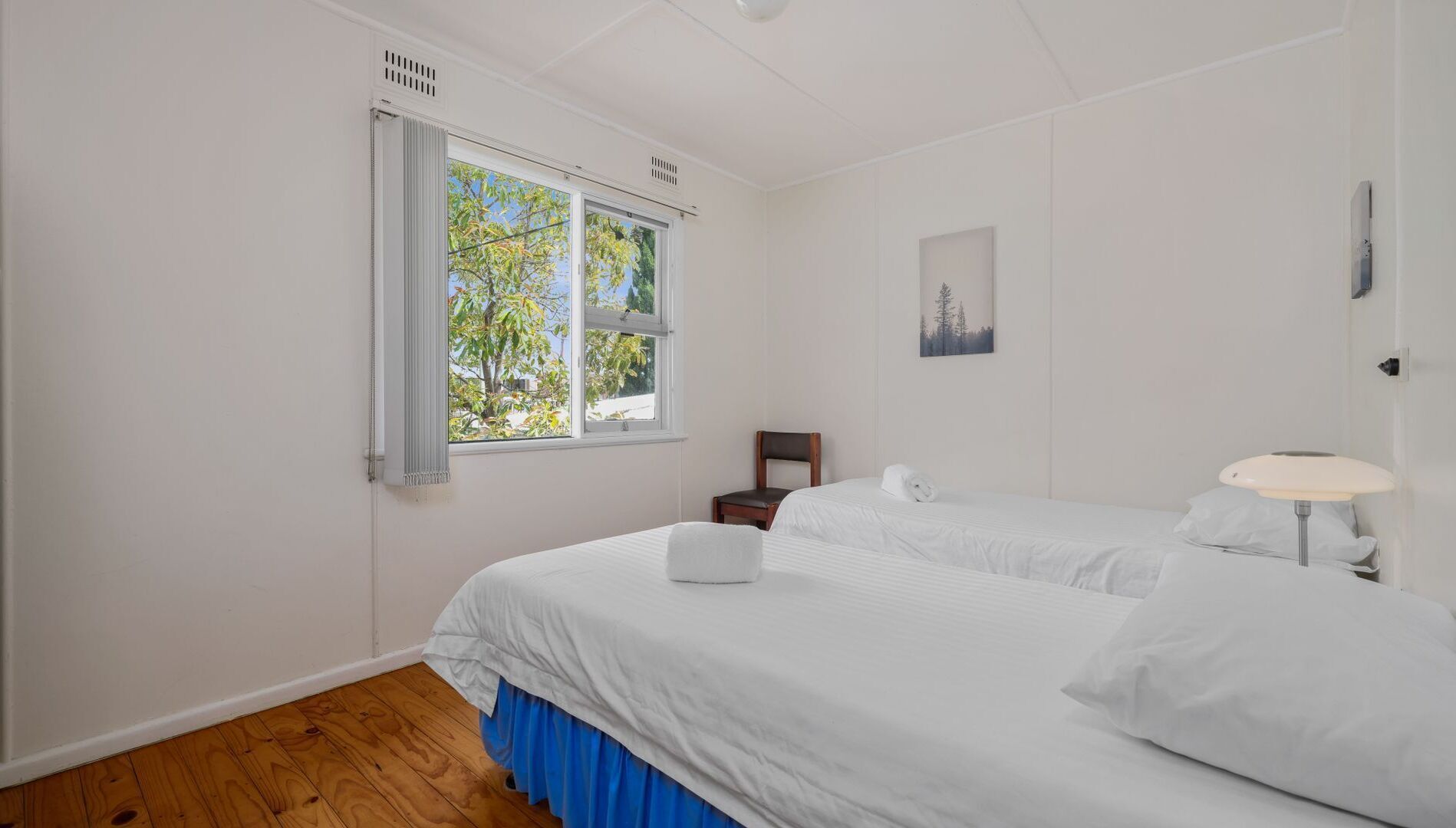 A bedroom with two beds, a window with a view of trees, a picture on the wall, and a bedside table with a lamp.