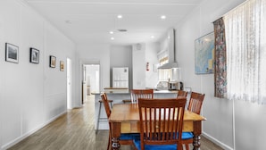 A dining area with a wooden table and chairs, a kitchen in the background, and a wall with framed pictures.