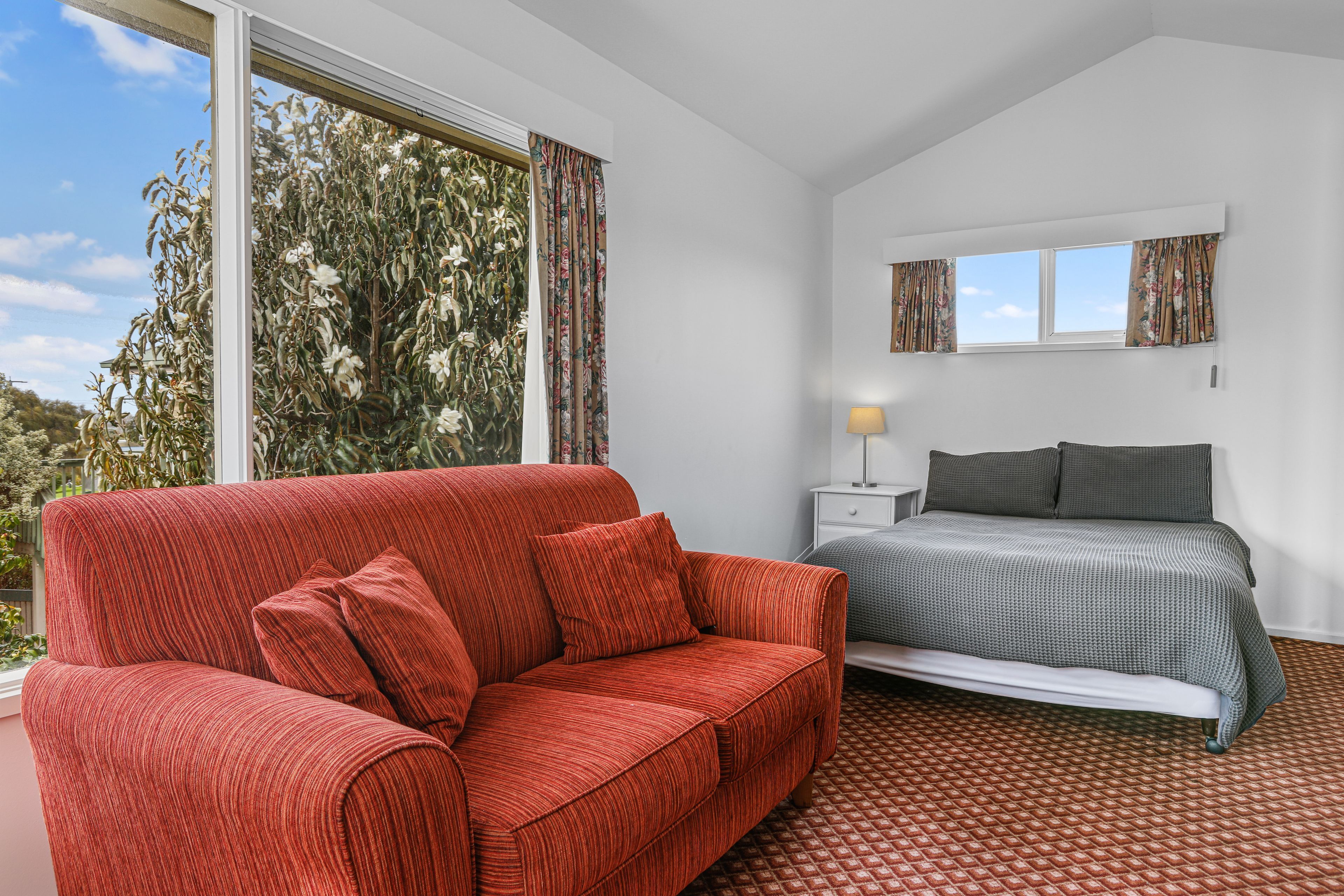 Une chambre avec un canapé rouge, un lit recouvert d’une couvre-lit à motifs, une table de chevet avec une lampe et une fenêtre avec des rideaux.