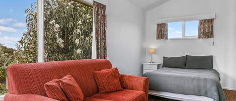 Une chambre avec un canapé rouge, un lit recouvert d’une couvre-lit à motifs, une table de chevet avec une lampe et une fenêtre avec des rideaux.