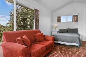 A bedroom with a red sofa, a bed with a patterned bedspread, a nightstand with a lamp, and a window with curtains.