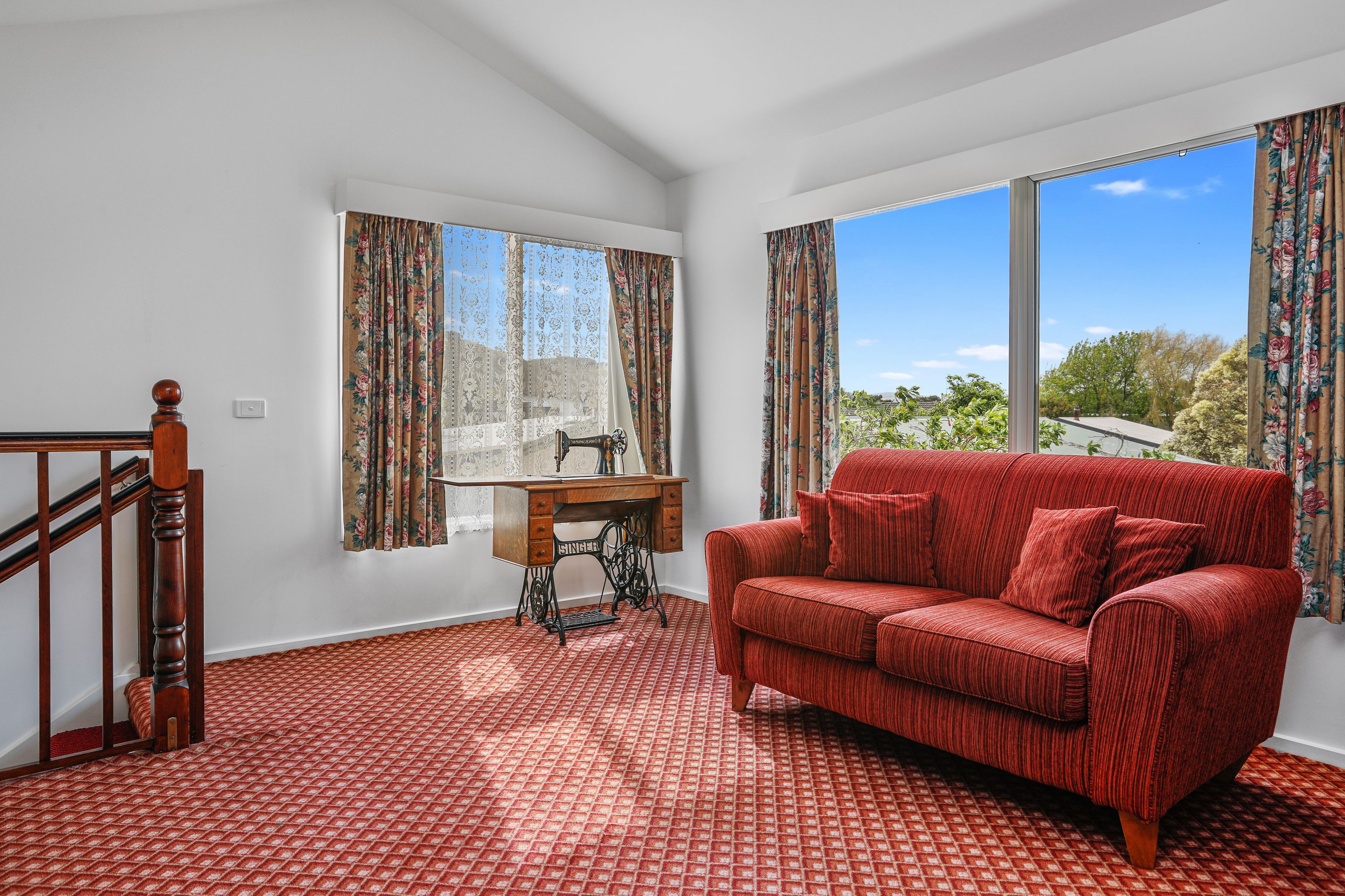 A room with a red patterned sofa, a wooden table with a sewing machine, and a window with floral curtains.