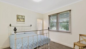 Ein Schlafzimmer mit einem Metallbettgestell, einem Einzelbett mit einem blauen, gemusterten Überwurf, einem Holzstuhl, einem Fenster mit Jalousien und einer Tür zu einem weiteren Raum.