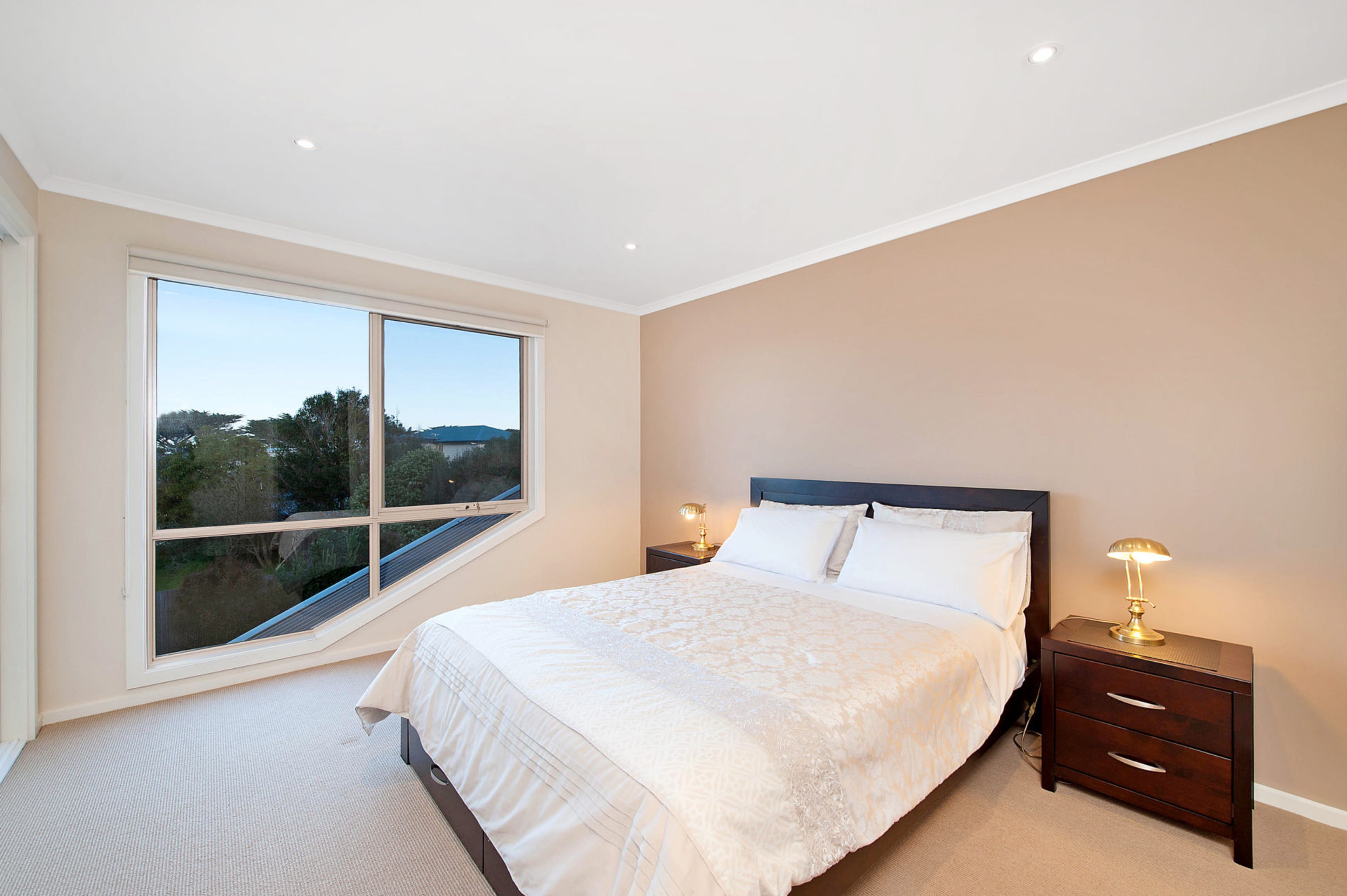A bedroom with a large window, a bed with white linens, two bedside tables with lamps, and a view of the outdoors.