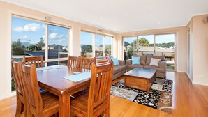 A spacious living area with a dining table, chairs, a sofa, and a coffee table. Large windows provide a view of the outdoors.