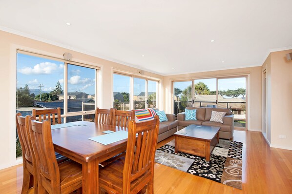 A spacious living area with a dining table, chairs, a sofa, and a coffee table. Large windows provide a view of the outdoors.
