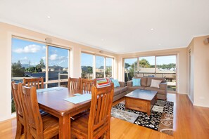 A spacious living area with a dining table, chairs, a sofa, and a coffee table. Large windows provide a view of the outdoors.