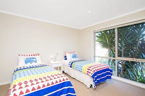 A bedroom with two beds, a window with a view of palm trees, and a white nightstand with a lamp. - Rose The Perfect Beach Escape Beachfront (Apollo Bay)