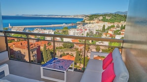 Un balcon avec vue sur la mer et les bâtiments, un canapé blanc avec des coussins rouges et une table à motifs.