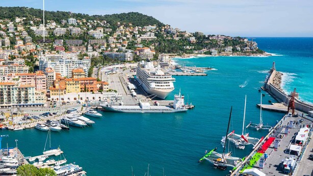 Un paysage urbain côtier avec une marina, des immeubles d’habitation et un grand navire de croisière amarré au port.