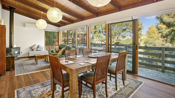 A spacious dining area with a wooden table and chairs, large windows, and a view of the outdoors. - Apollo Bay People N Paws (Apollo Bay)