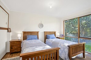 A bedroom with two wooden beds, a bedside table with a lamp, a wall-mounted mirror, and a large window with a view of trees. - Apollo Bay People N Paws (Apollo Bay)