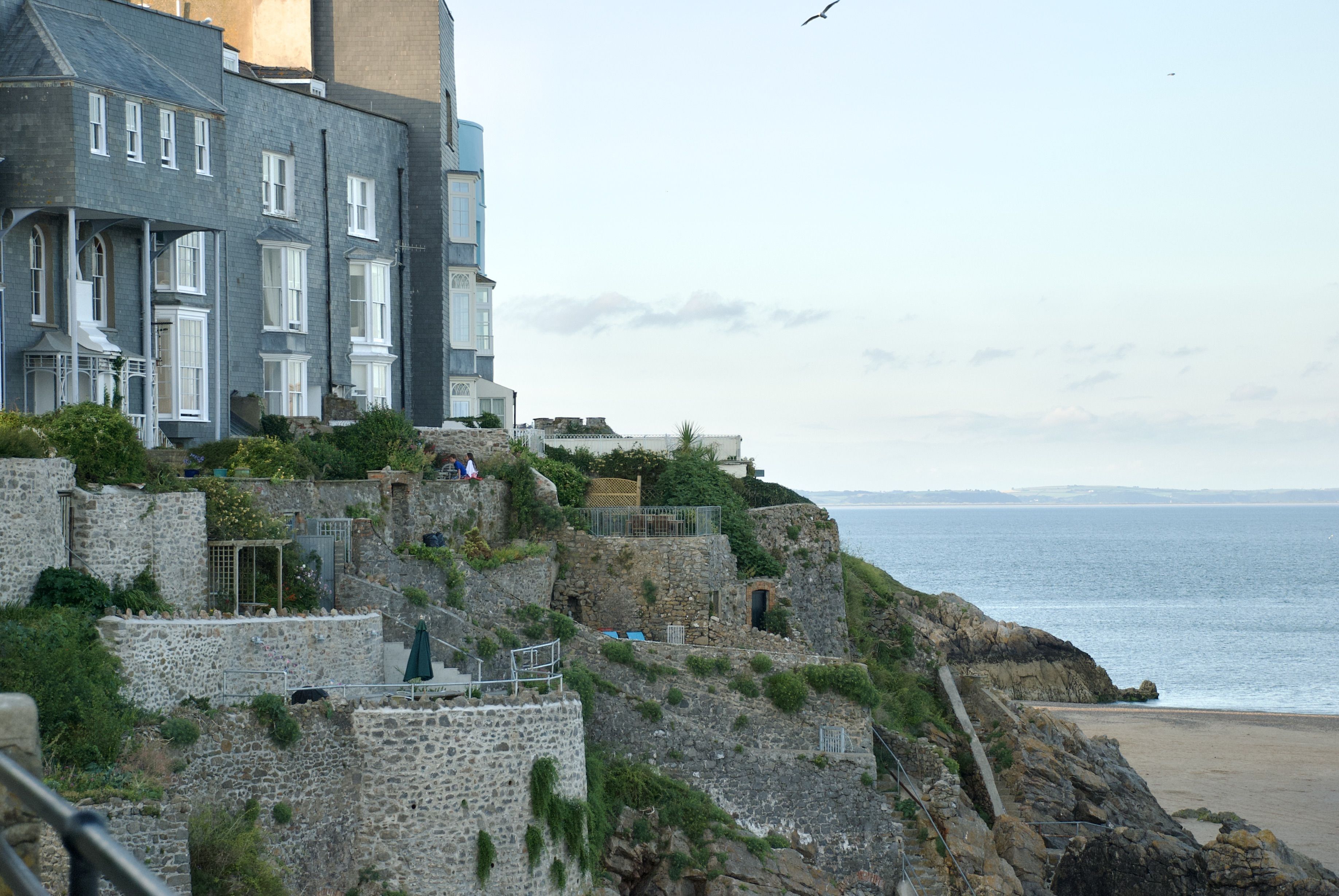 Um edifício histórico de pedra, com vários andares, uma escadaria que leva aos pisos superiores e vista para o mar.