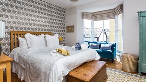 A bedroom with a bed, a blue sofa, a wooden bench, a patterned rug, and a window with curtains. - High House (6), Tenby , Pembrokeshire (Tenby)