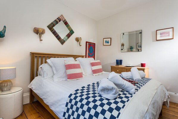 A neatly made bed with a checkered blanket, a wooden headboard, and striped pillows. There is a nightstand with a lamp and a small decorative item. A mirror and framed artwork are on the wall.