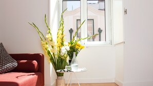 A room with a red sofa, a small table with a vase of flowers, and a window with a view of buildings. - Arthistorian's Residence ★ central & sunny (Lisboa)