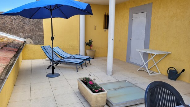 Un balcone con due poltrone a righe, un ombrellone blu, una pianta in vaso, un tavolino pieghevole bianco e un annaffiatoio.