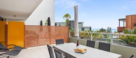 A modern outdoor dining area with a table and chairs, a wooden fence, and a view of the sea.