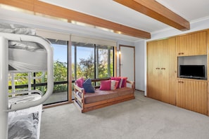 A room with a bunk bed, a sofa with cushions, a TV on a wooden cabinet, and a large window with a view of trees. - Retro Retreat (Lorne)