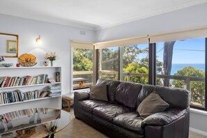 A living room with a leather sofa, a glass coffee table, and a bookshelf filled with books. There is a window with a view of trees and the sea.
