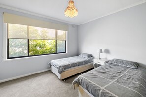 A bedroom with two single beds, a window with blinds, a bedside table with a lamp, and a hanging light fixture. - Retro Retreat (Lorne)