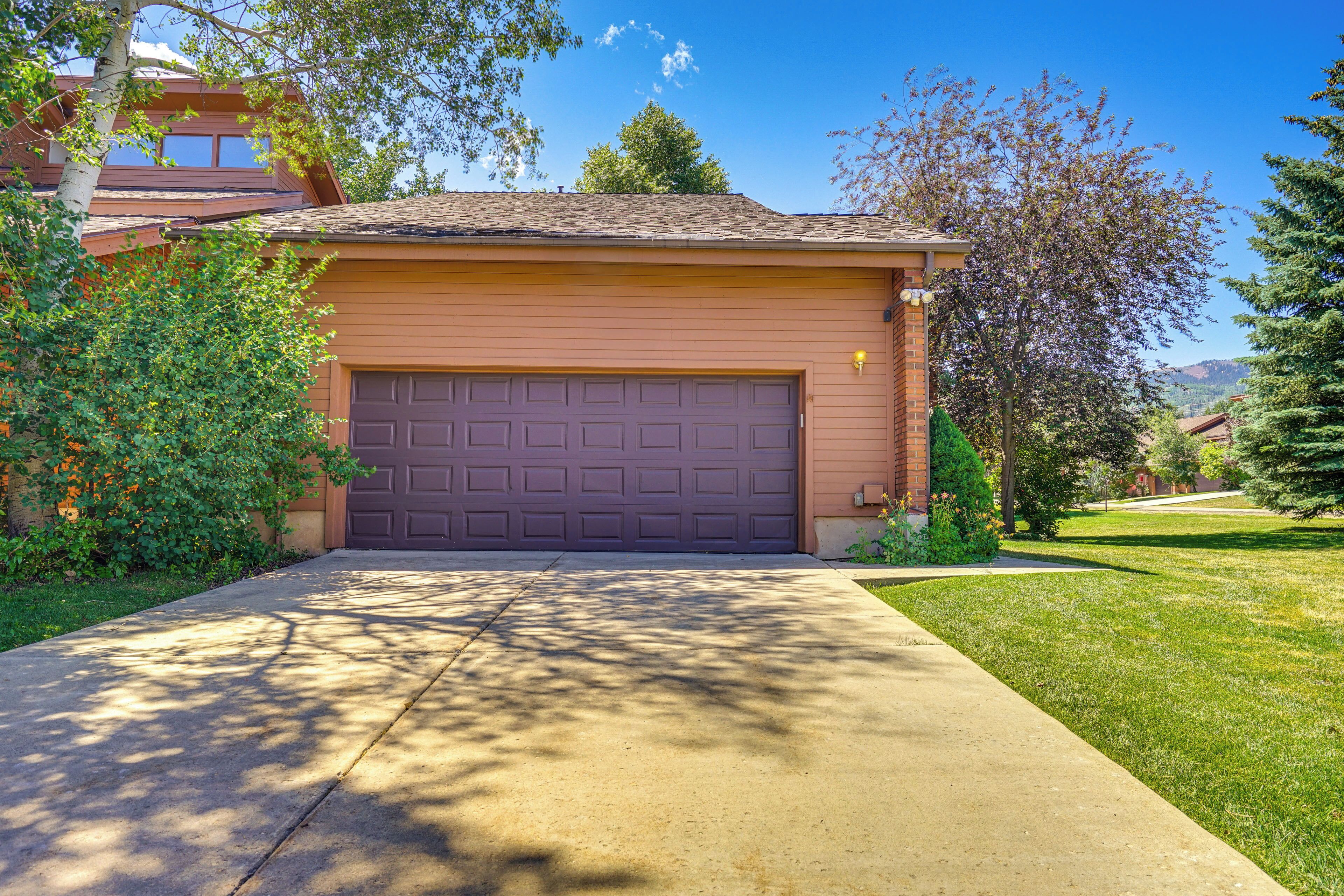 A residential garage with a brown door, surrounded by greenery and a clear sky.