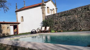 Zona de piscina con un muro de piedra, un edificio blanco de techo rojo y mobiliario exterior.