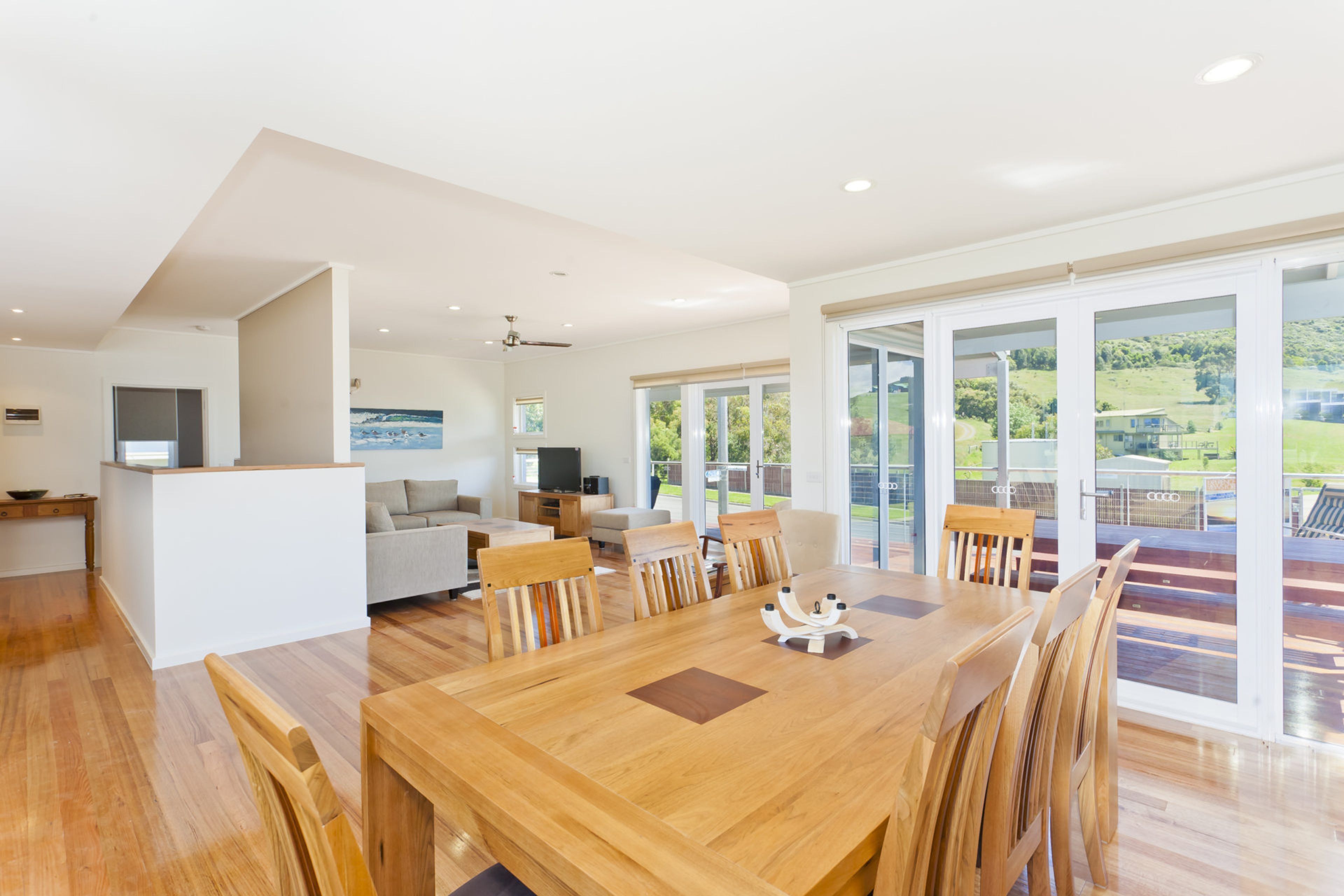 A spacious dining area with a wooden table and chairs, a kitchen area with a television, and large sliding glass doors leading to a balcony with a view of greenery.