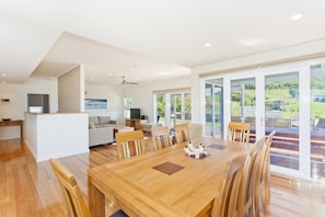 A spacious dining area with a wooden table and chairs, a kitchen area with a television, and large sliding glass doors leading to a balcony with a view of greenery.