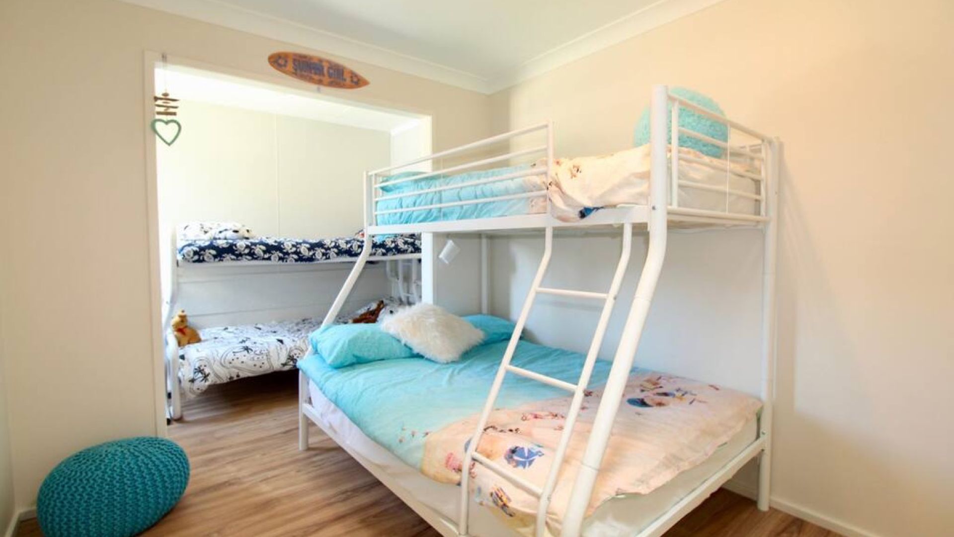 A bunk bed room with a white ladder, a blue and floral bedspread, and a teal pouf on the floor.