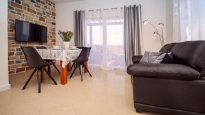 Uma sala de estar com sofá, mesa de jantar com cadeiras, televisão de tela plana fixada em uma parede de tijolos e vista pela janela com cortinas.