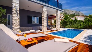 Uma área de piscina moderna ao ar livre, com espreguiçadeiras e uma mesa de jantar, cercada por pilares de pedra e vista para uma montanha.