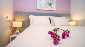 Uma cama bem arrumada com lençóis e almofadas brancas, um abajur e um quadro de flores na parede.