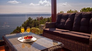 Um sofá de vime com almofadas, uma mesa de vidro com uma tigela de frutas e dois copos de suco de laranja, e vista para o mar.