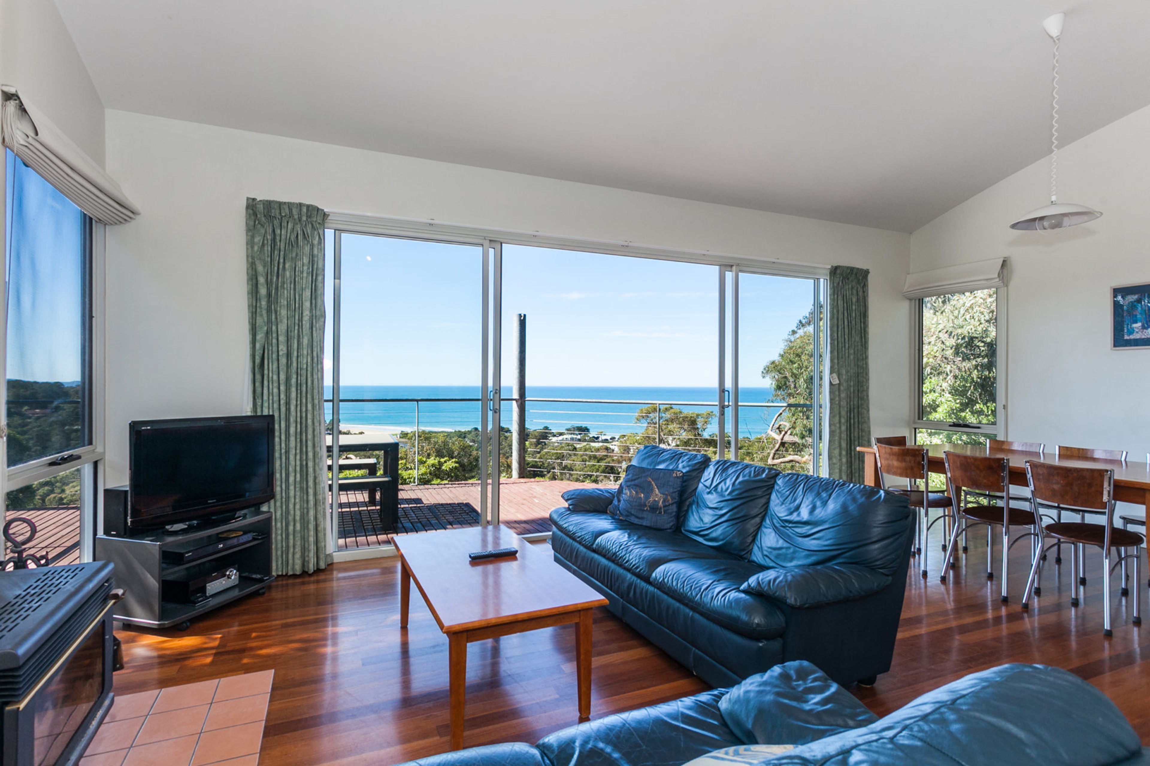 A living room with a sofa, television, and a table. There is a view of the ocean through large windows.