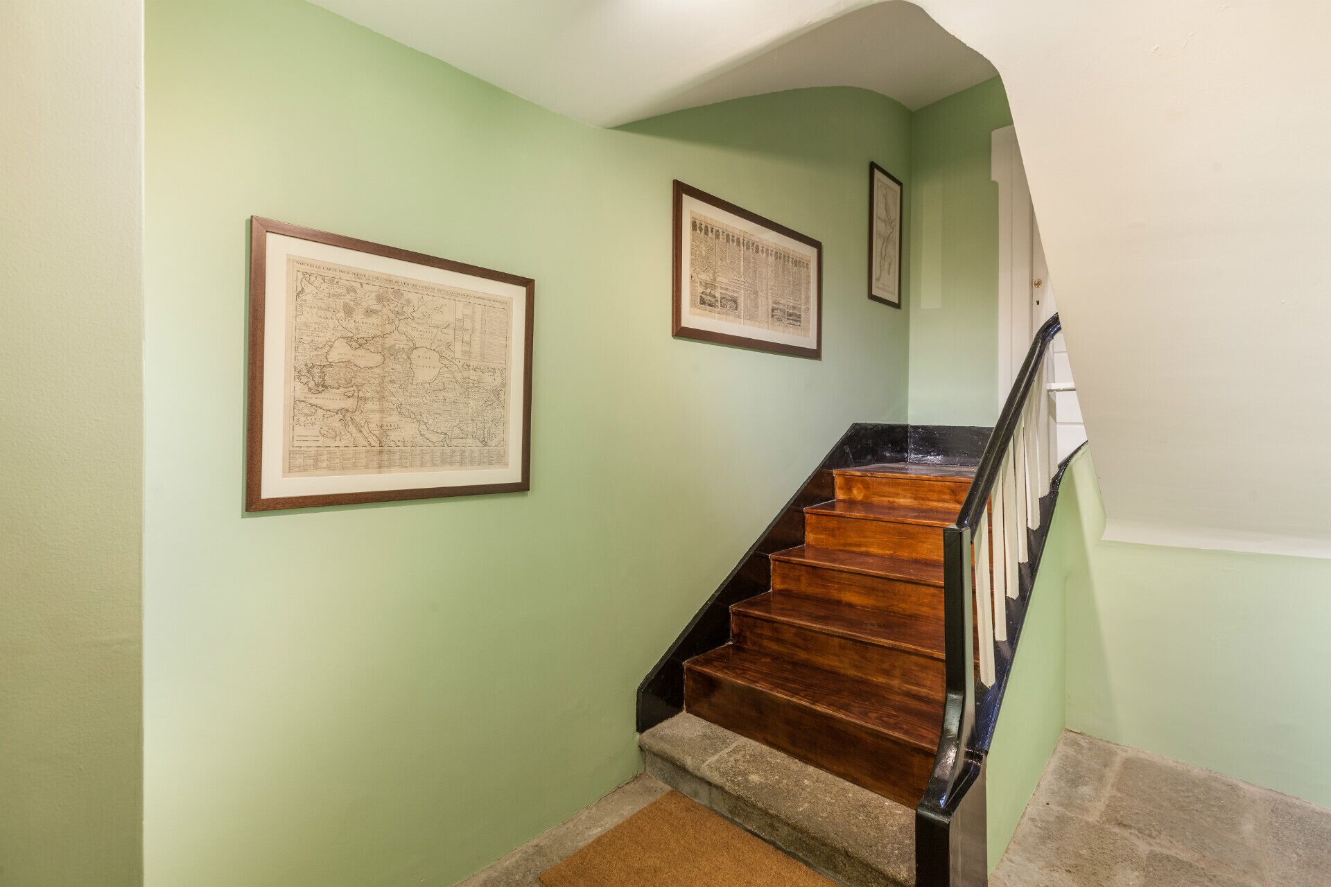 A staircase with wooden steps and black railings, flanked by light green walls and framed historical maps.