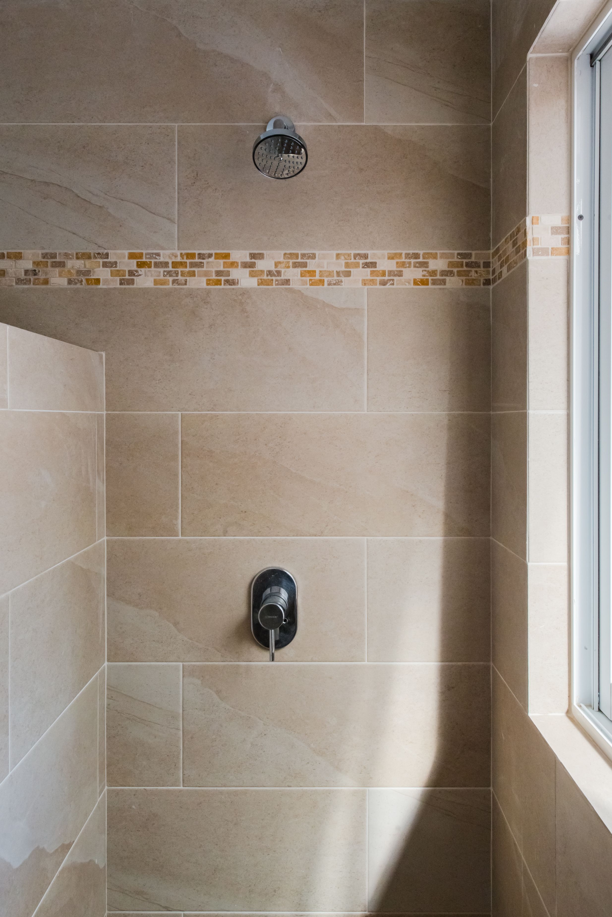 Un área de ducha con azulejos color crema, un ventanal pequeño y un cabezal de ducha.
