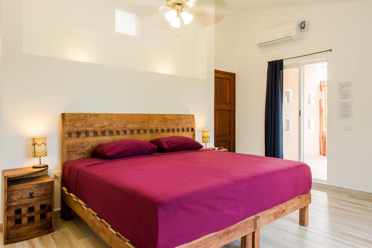 Un dormitorio con estructura de cama de madera, colcha roja, dos mesitas de noche con lámparas, un ventilador de techo y una puerta corrediza de vidrio que da a un espacio exterior.