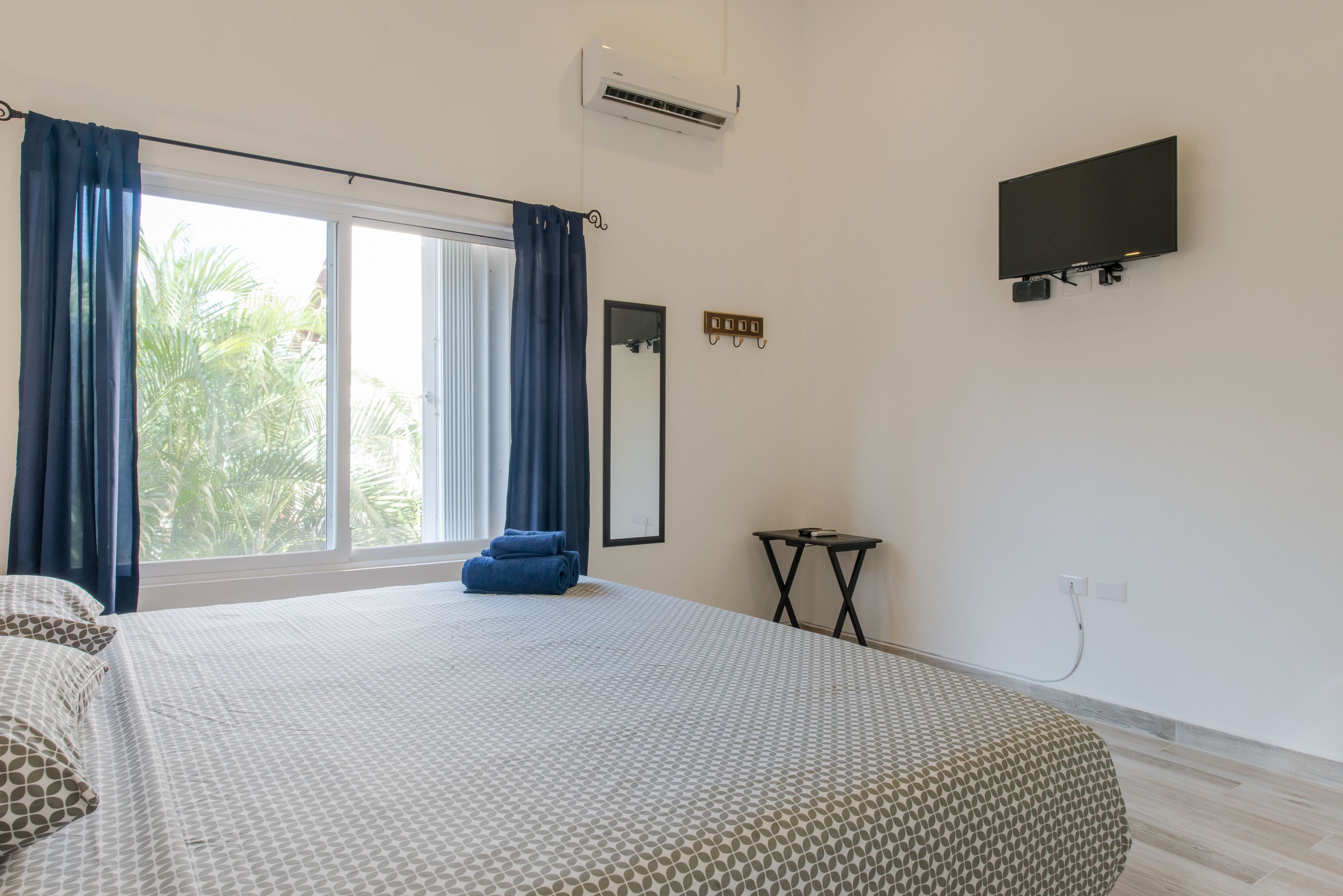 A hotel room with a large bed, a TV mounted on the wall, a small table, and a window with curtains.