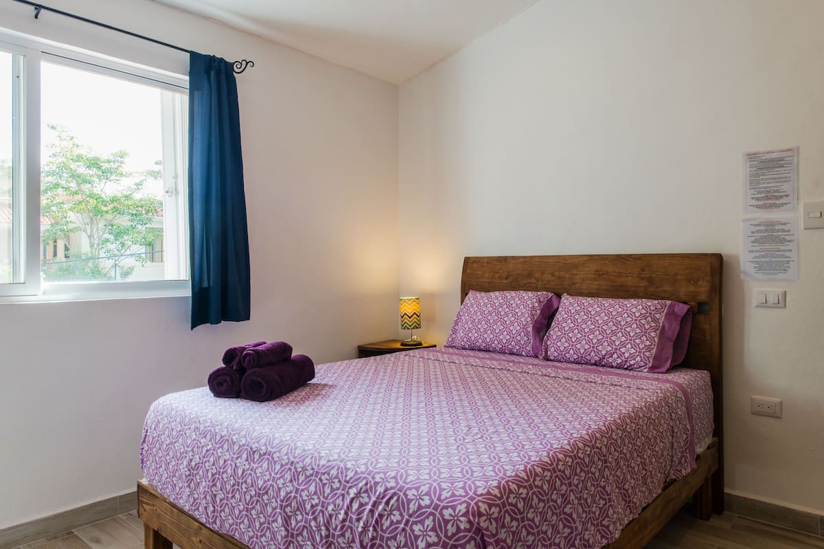 Un dormitorio con una cama, una ventana con cortinas, una mesita de noche con una lámpara y una pared con papeles.