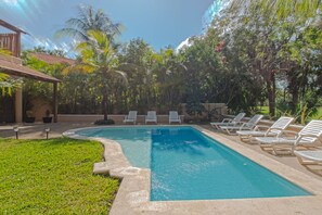 A rectangular swimming pool with a tiled deck, surrounded by white lounge chairs and lush greenery, including palm trees and various shrubs. - Villas Picalu Studios & Suites (Puerto Aventuras)