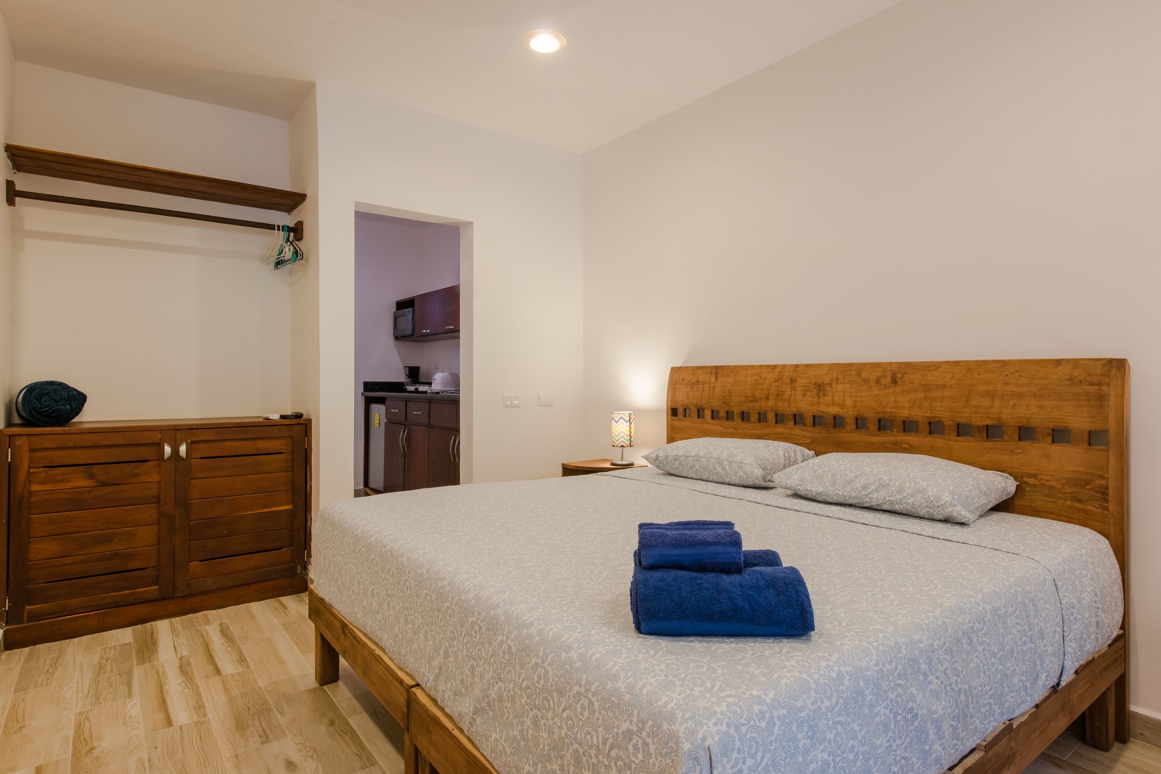 A bedroom with a wooden bed, a wooden wardrobe, and a small kitchenette in the background.