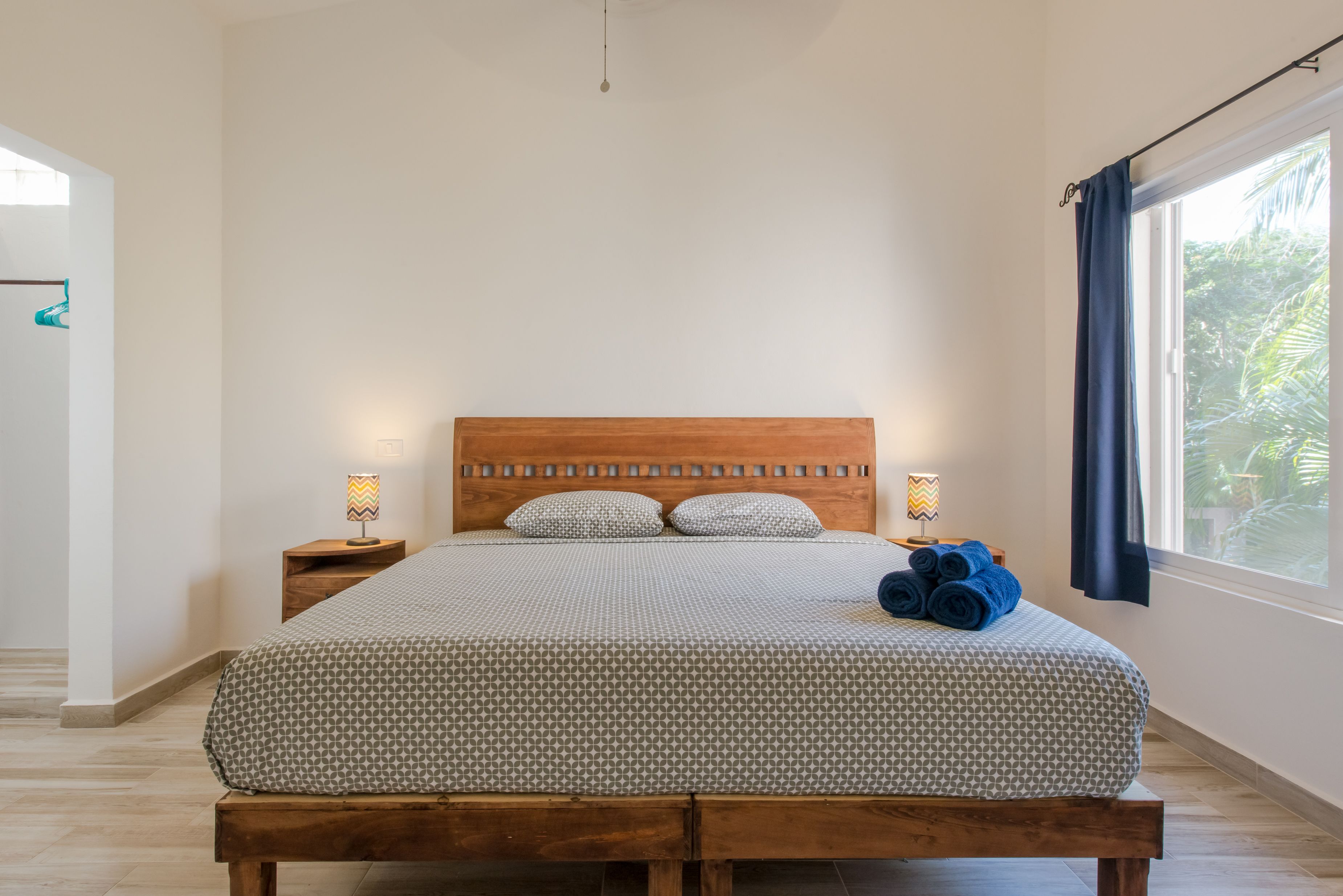 A bedroom with a wooden bed, two bedside tables, a window with curtains, and a view of greenery outside.