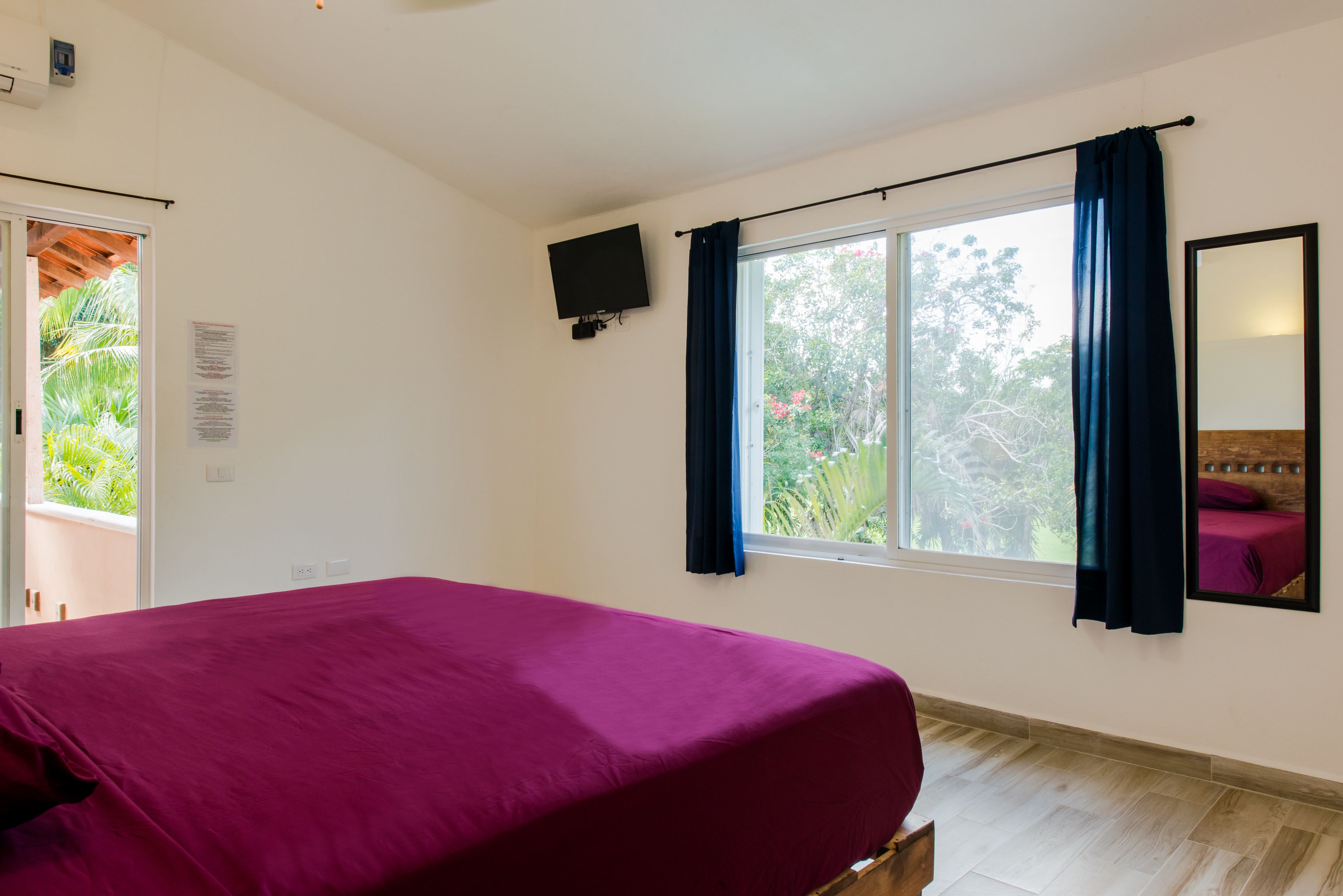 Ein Schlafzimmer mit einem Bett, einem Vorhangfenster, einem an der Wand montierten Fernseher und einer Tür, die nach draußen führt.