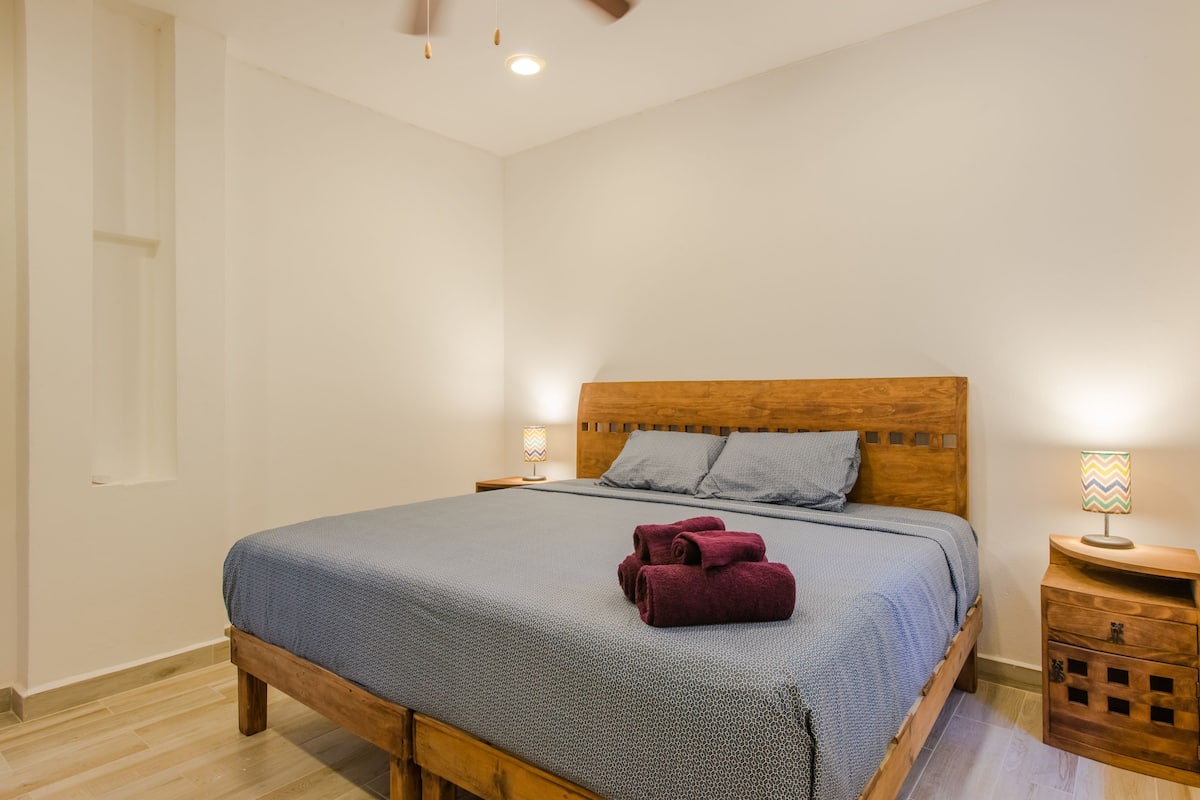 Una cama bien hecha con cabecera de madera, dos almohadas y una colcha azul. Hay dos mesitas de noche con lámparas y un pequeño tocador de madera con una lámpara.